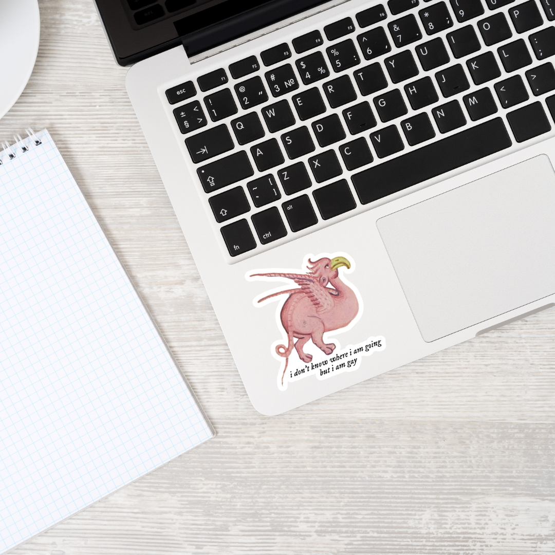 Matte vinyl sticker featuring a pink griffin with yellow beak and wings, curled tail, and the text ‘i don't know where i am going but i am gay’ displayed on a laptop, showcasing its whimsical design and matte finish