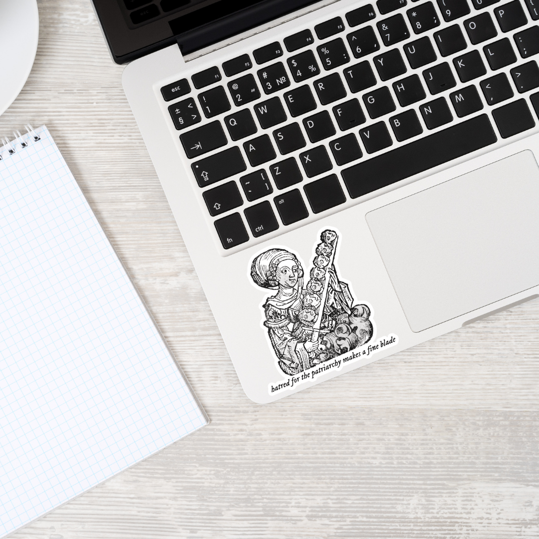 Medieval-style black and white sticker of a woman wielding a sword of severed heads with the text: ‘Hatred for the patriarchy makes a fine blade'. The sticker is displayed on a laptop, showcasing bold feminist and gothic design