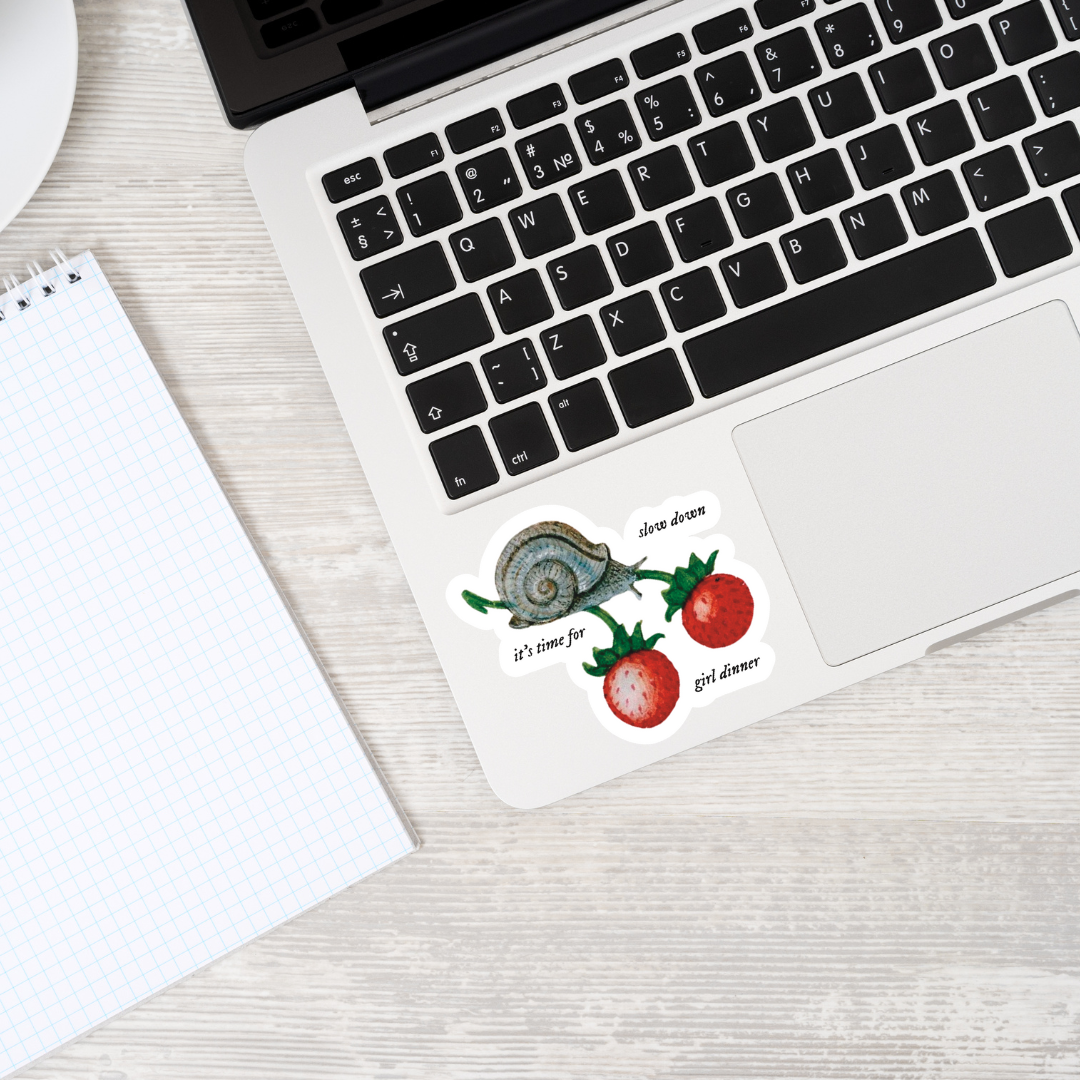 Matte vinyl sticker with the text ‘Slow Down, It’s Time for Girl Dinner’ in medieval manuscript-inspired font displayed on a laptop, showcasing its whimsical design and matte finish.